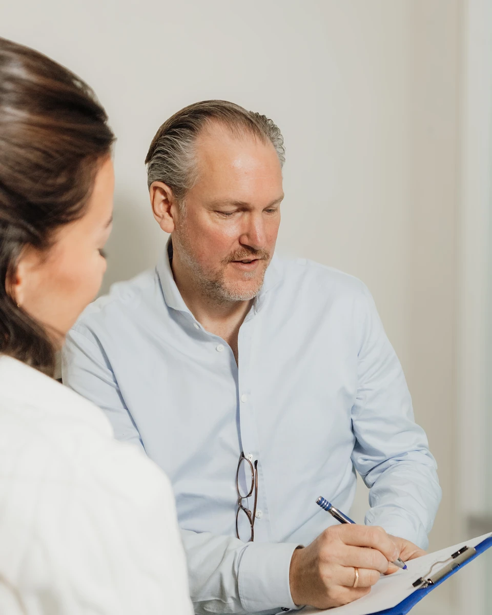 Dr. Meschede im Erstgespräch mit Klemmbrett, die Patientin ist von hinten zu sehen